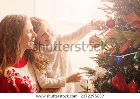 Happy woman in red sweater and child in white sweater hang Christmas ball on the Christmas tree together. Mom and daughter