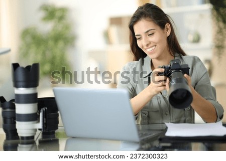 Happy photographer checking work on laptop holding camera at home