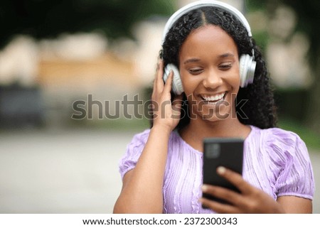 Front view portrait of a happy black woman listening to music wearing headphone checking phone in the street