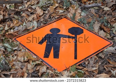 An orange traffic sign with a person holding a stop sign. The sign is on the ground surrounded by fall leaves.