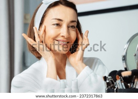 Smiling close-up portrait of an adult woman housecoat garment in bathroom touching face with hands and posing in front of makeup mirror Royalty-Free Stock Photo #2372317863