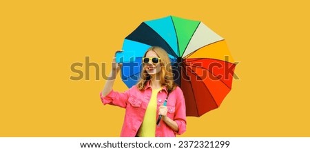 Portrait of stylish happy smiling young woman taking selfie with mobile phone holds colorful umbrella wearing pink jacket on yellow studio background