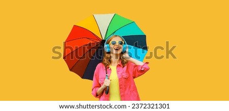Happy cheerful laughing young woman with colorful umbrella listening to music in headphones wearing pink jacket on yellow studio background