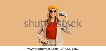 Fashionable autumn color style outfit, stylish young woman photographer with film camera wearing orange french beret hat, jacket and round sunglasses on brown studio background