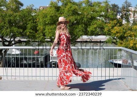 Pretty young blonde woman with straw hat on her head. She is dressed in a white dress with red print and is having fun doing different poses. The woman is on holiday in Spain