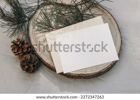 Christmas stationery. Blank greeting card, invitation mockup on cut wooden round board. Green pine tree branch in sunlight. Grey linen tablecloth. Festive winter still life template, flat lay, top Royalty-Free Stock Photo #2372347263