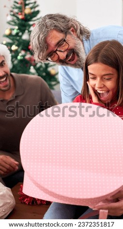 Vertical. Surprised girl opening Christmas presents at home. Happy and excited family wearing Santa Claus hat gathered together for gift exchange. Positive and joyful domestic relationships in winter
