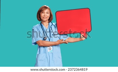 Happy smiling asian nurse holding red speech bubble sign of empty space for text. Smiling healthcare specialist presenting thought bubble cardboard, isolated over blue studio background