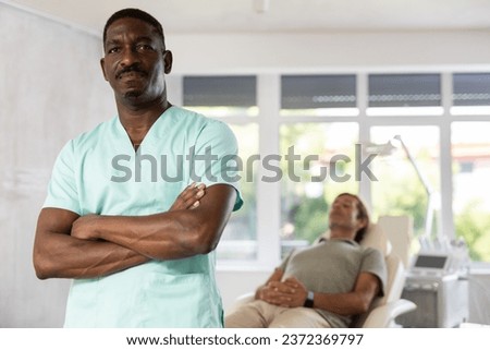 Experienced African male doctor is standing in office, behind him unrecognizable patient is waiting for consultation. Advertising poster banner of medical clinic, cosmetology office, hospital