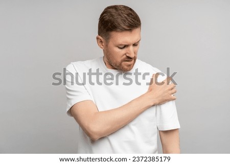Man holds on to sore shoulder. Frowning dissatisfiedly, middle aged guy experiences throbbing pain in ligaments, suffers discomfort, squeezes sore spot and sighs. Studio portrait isolated on gray. Royalty-Free Stock Photo #2372385179