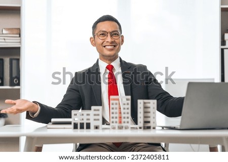 Agent presents contracts for property purchase or lease. Businessman asian indian people showcases miniature model home, signifying secure property insurance. Desk scene in a home sales office.