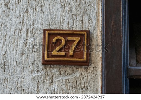 Wooden brown plate board, with engraved number 27. On the white wall background. Part of the house , building architecture. Twenty seven.