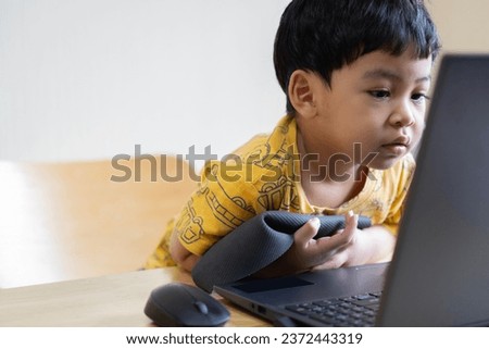 Cheerful little Asian kid boy 3s wearing casual shirt looking on laptop pc computer on light biege color wall background. Childhood lifestyle concept