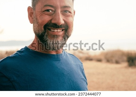 Portrait outdoor of adult mature cheerful man smiling at the camera. Bearded caucasian male people with blue t-shirt with happy serene expression on face. People in outdoors leisure activity alone. Royalty-Free Stock Photo #2372444077