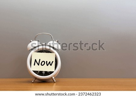 Paper note word Now on alarm clock wood desk white background. 
Sticker note paper message Now and watch on wooden table for copy space.