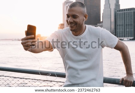 Young man with headphones taking selfie against background of river and city