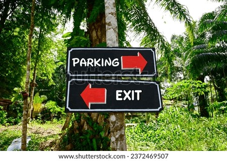 Two black wooden signs with white frames, red arrows pointing in opposite directions with white text The parking lot has an arrow pointing to the right, the exit has an arrow pointing to the left.