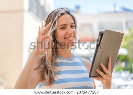 Young pretty woman holding a tablet at outdoors showing ok sign with fingers