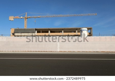 Fence consisting of plastic sheeting at the roadside outside the construction site. Building under construction with tower crane above on behind,road in front. Background for copy space. 