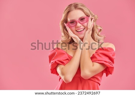 Young beauty. A cute attractive girl with blond hair poses in a pink dress and pink glasses and smiles. Pink studio background with copy space. Femininity, beauty and fashion.