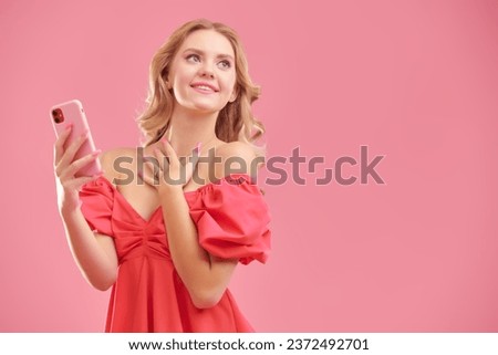 A young fashionista girl with beautiful curly blond hair, dressed in an elegant pink dress, chats emotionally on the phone. Studio pink background with copy space.
