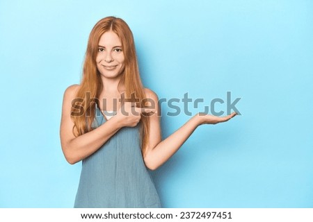 Redhead young woman on blue background excited holding a copy space on palm.