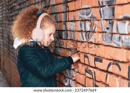 Girl hand in the gap of the wall. Teenager reaches out to wall hole. Child by the brick wall. The concept of silence, mystery, suspense, interest Depressed girl. Gloomy child on brick background.  Royalty-Free Stock Photo #2372497669
