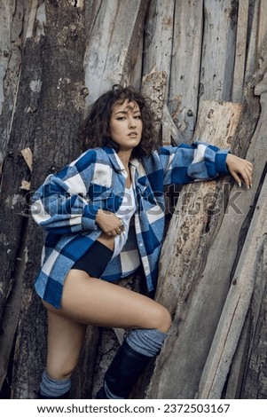 Portrait of a beautiful young fashionable woman in a plaid shirt stands against the background of an old barn house in the village in summer