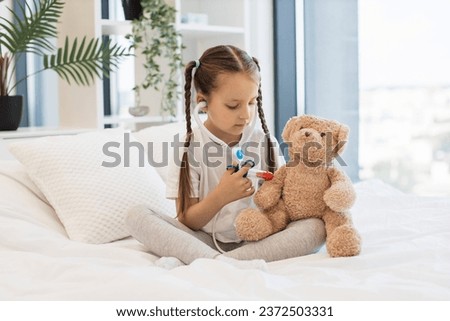 Gentle caucasian girl dream of becoming doctor and training to giving vaccinations to favorite soft toys. Pretty child with stethoscope and small toy syringe doing injecting to teddy bear.