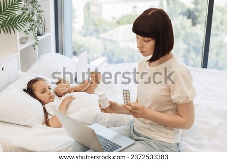 Focused mother using wireless laptop for video chatting with doctor while sitting on bed near pretty unwell daughter. Caucasian woman showing various pills in hands while looking on computer screen. Royalty-Free Stock Photo #2372503383