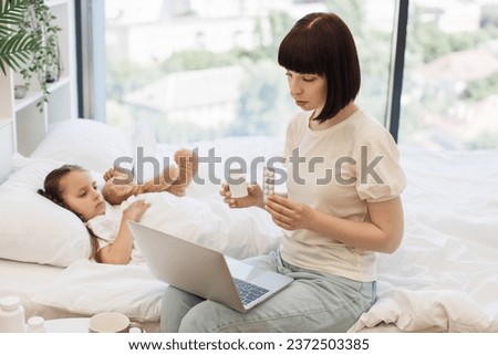 Focused mother using wireless laptop for video chatting with doctor while sitting on bed near pretty unwell daughter. Caucasian woman showing various pills in hands while looking on computer screen. Royalty-Free Stock Photo #2372503385