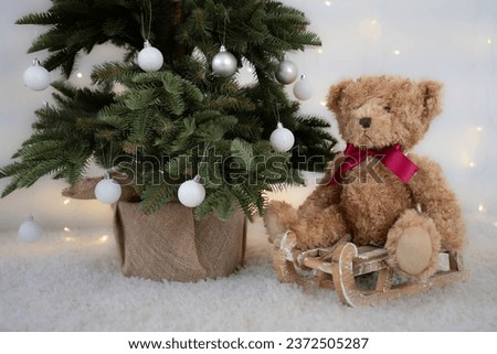 Brown Teddy Bear with red bow, who is sitting on a wooden sledge, next to a Christmas tree with white decorations and natural basket. White carpet and Christmas lights in the background. Advent theme.