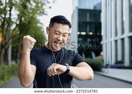 Close-up photo. A young Asian man does sports, runs on the city street in headphones, looks at the result on a smart watch, rejoices, showing a victory gesture with his hand.