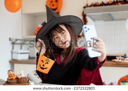 An adorable and joyful young Asian girl in a Halloween costume is taking selfies or her video with a smartphone in the kitchen, celebrating Halloween at home. Halloween season concept
