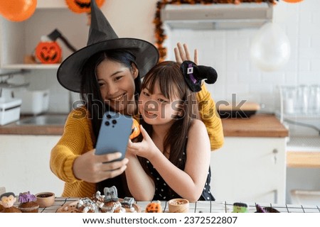 A cute and happy young Asian girl in a Halloween costume is having fun, taking selfies or taking video with her mom while making Halloween cupcakes and celebrating Halloween at home together.