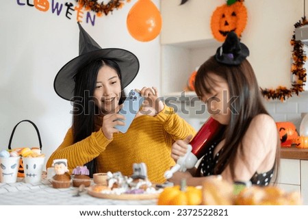 A kind and beautiful young Asian mom in a witch hat is having fun, enjoys making Halloween cupcakes with her young daughter in the kitchen, and celebrates Halloween at home together.