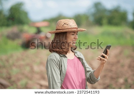 Young woman standing on field in sunset while tractor baling in background, Farmer or agrarian with smart phone looking on growing efficiency and crop yield, Smart agriculture and automation concept.