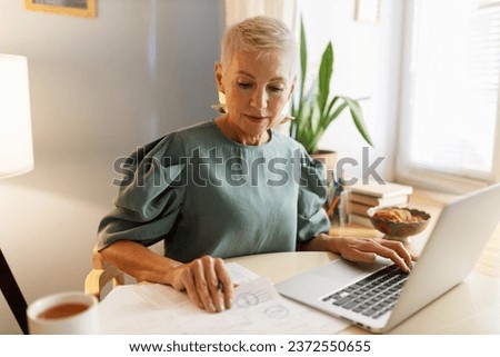 Beautiful elegant mature concentrated stylish female director general working with papers and documents in front of laptop, at his home office or cabinet next to window in stylish dress