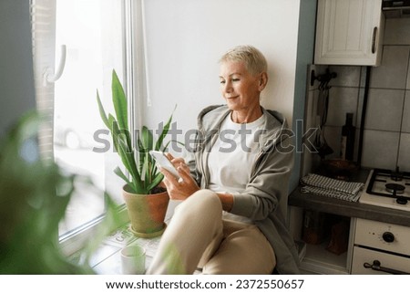 Charming stylish sporty female of 50 sitting on windowsill at kitchen browsing social media on smartphone, surfing mobile internet, texting, commenting pictures after jogging outside in morning