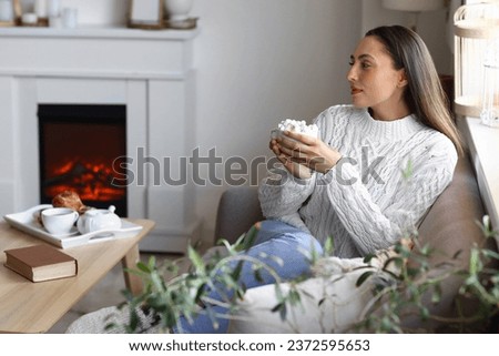 Young woman drinking cocoa with marshmallows near fireplace at home