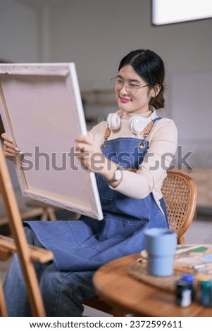 Young asian female artist wear headphone in her neck and looking artwork on canvas with happiness.
