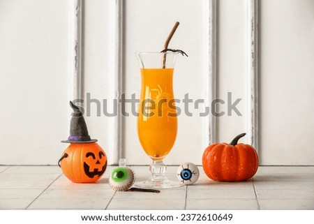 Glass of delicious orange cocktail, pumpkin and eyes for Halloween celebration on light background