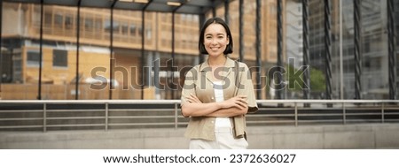 Portrait of beautiful young woman in casual clothes, smiling, posing outdoors on an empty street near glass building.