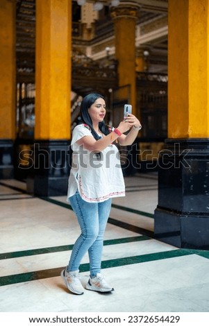 Happy young mexican woman tourist in embroidered top looking at screen of smartphone while standing in Postal palace in downtown Mexico City and browsing pictures in light against blurred interior