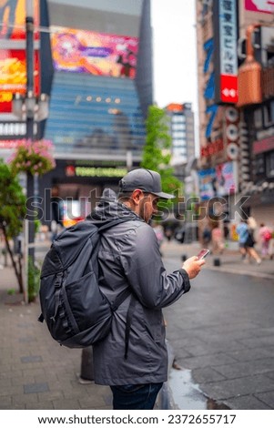 Side view of concentrated young Hispanic male tourist in warm clothes and cap with backpack taking photo on buildings on smartphone while standing in Akihabara neighborhood in Tokyo, Japan