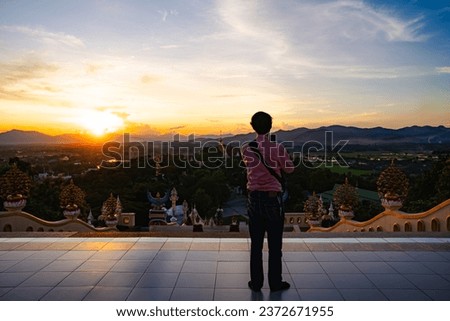 A view of mountains, houses, and temples from a high angle, with people standing and taking pictures from smartphones. During the time the sun is setting.