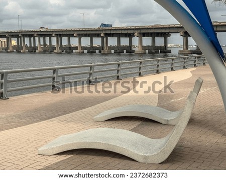 Pair of sculpturesque concrete recliners along a downtown riverwalk, with view of trucks on highway bridge across the Manatee River, on a cloudy afternoon in Bradenton, Florida, along the Gulf Coast