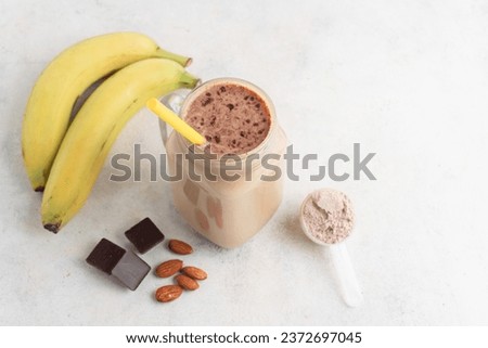Glass jar of protein milkshake drink or smoothie and whey protein powder in measuring spoon, bananas, chocolate cubes, almond nuts on white background. sport nutrition, bodybuilding food supplements. Royalty-Free Stock Photo #2372697045