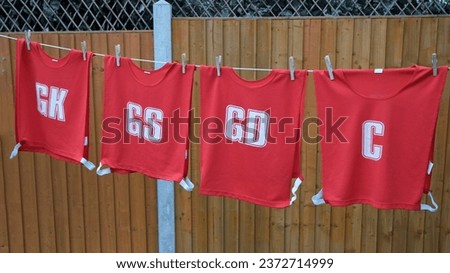 Red sports or netball bibs hanging with pegs on a washing line to dry. Team position in letters on bib. Brown orange panel fence in background. Overcast day outdoors. Close up Royalty-Free Stock Photo #2372714999