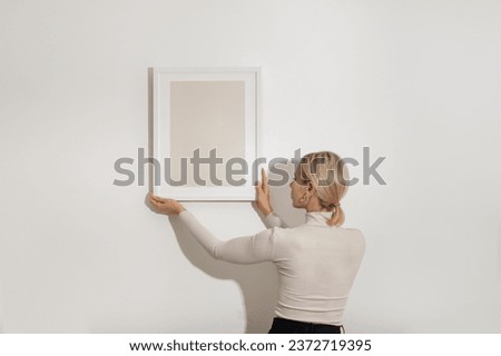 Blond young woman hanging a blank white wooden frame on the white wall. Minimalistic lifestyle, copy space for your text.
 